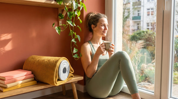 Frau in Wellness-Pause am Fenster mit Kräutertee, Faszienrolle in gelber Yogadecke